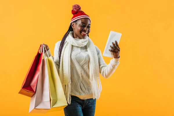 Mujer afroamericana en traje de invierno sosteniendo bolsas de compras y tabletas digitales aisladas en amarillo - foto de stock