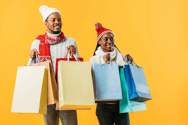 Casal afro-americano em roupa de inverno segurando sacos de compras e olhando para longe isolado no amarelo — Fotografia de Stock