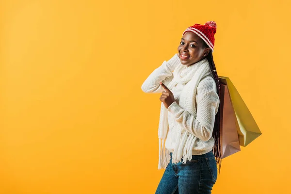 Side view of African American woman in winter outfit holding shopping bags and pointing with finger isolated on yellow — Stock Photo