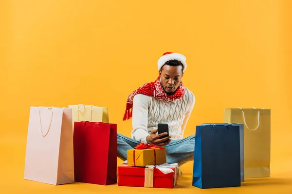 African American man in winter outfit sitting and taking pictures of presents isolated on yellow — Stock Photo
