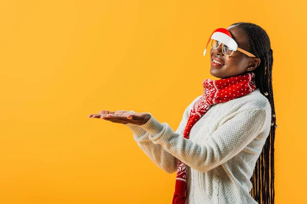 Vista lateral da mulher afro-americana em camisola de inverno e óculos de Natal de mãos dadas com palmas olhando para cima isolado no amarelo — Fotografia de Stock