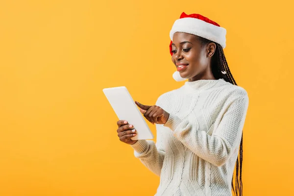 African American woman in winter sweater and Santa hat tapping on screen of digital tablet isolated on yellow — Stock Photo