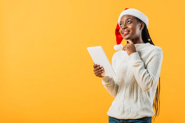 Dreamy African American woman in winter sweater and Santa hat holding digital tablet isolated on yellow — Stock Photo