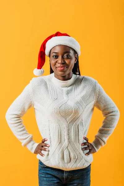 Donna afroamericana in maglione invernale e cappello di Babbo Natale che si tiene per mano sui fianchi e guarda la fotocamera isolata sul giallo — Foto stock