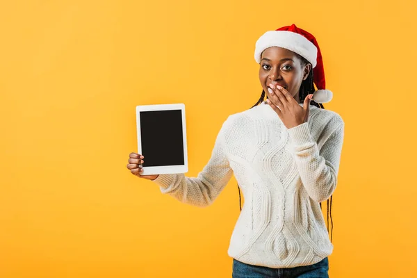 Mujer afroamericana en suéter de invierno y Santa hat sosteniendo tableta digital con pantalla en blanco y cubriendo la boca con la mano aislada en amarillo - foto de stock
