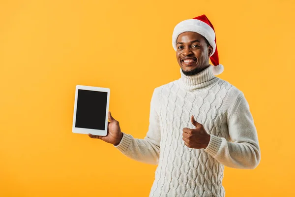 Hombre afroamericano en suéter de invierno y sombrero de Santa sosteniendo tableta digital con pantalla en blanco y dando pulgar hacia arriba aislado en amarillo - foto de stock