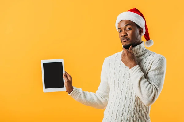 Hombre afroamericano en suéter de invierno y sombrero de Santa celebración tableta digital con pantalla en blanco y la barbilla acariciando aislado en amarillo - foto de stock