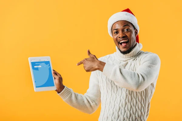KYIV, UCRAINA - 2 AGOSTO 2019: Uomo afroamericano in cappello da Babbo Natale puntato con il dito contro tablet digitale con app twitter isolata su giallo — Foto stock