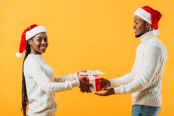 Vista lateral de pareja afroamericana en sombreros de Santa sosteniendo caja de regalo aislada en amarillo - foto de stock