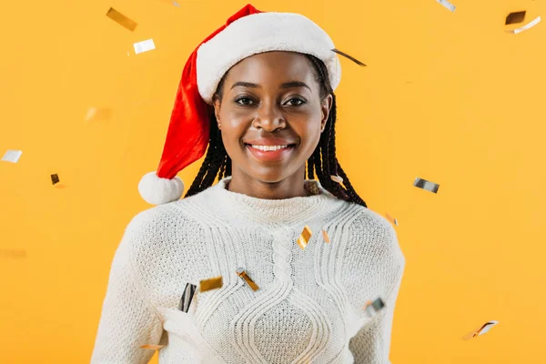 Mulher afro-americana em chapéu de Santa sorrindo e olhando para a câmera no fundo amarelo com confete — Fotografia de Stock
