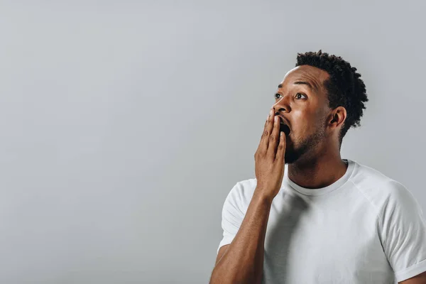 Homme afro-américain choqué en blanc T-short couvrant la bouche avec la main et regardant la caméra isolée sur gris — Photo de stock