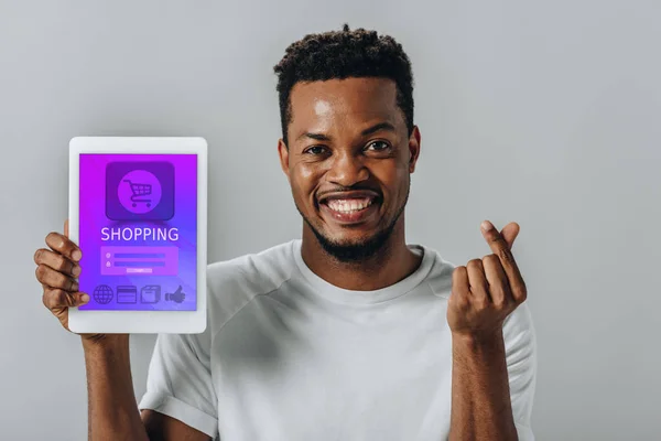 African American man holding digital tablet with shopping application and showing money gesture isolated on grey — Stock Photo