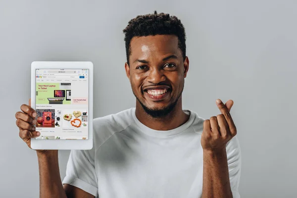 KYIV, UKRAINE - AUGUST 2, 2019: African American man holding digital tablet with ebay website and showing money gesture isolated on grey — Stock Photo