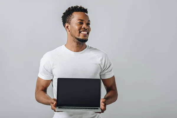 Hombre afroamericano sonriendo y mostrando portátil con pantalla en blanco y mirando hacia otro lado aislado en gris — Stock Photo