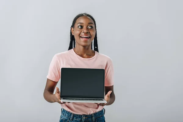 Mujer afroamericana emocionada sosteniendo el ordenador portátil con pantalla en blanco y mirando hacia otro lado aislado en gris - foto de stock