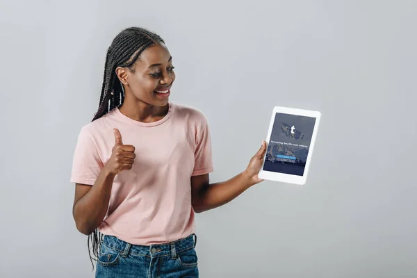 KYIV, UKRAINE - AUGUST 2, 2019: African American woman holding digital tablet with tumblr app and showing thumb up isolated on grey — Stock Photo