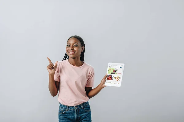 KYIV, UKRAINE - 2 AOÛT 2019 : Femme afro-américaine tenant une tablette numérique avec site Web ebay et pointant du doigt isolé sur gris — Photo de stock