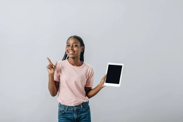 Афроамериканка держит цифровой планшет с чистым экраном и указывает пальцем, изолированным на сером — стоковое фото