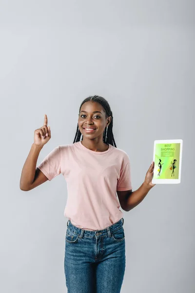 Mulher afro-americana segurando tablet digital com aplicação de compras e mostrando sinal de ideia isolado no cinza — Fotografia de Stock