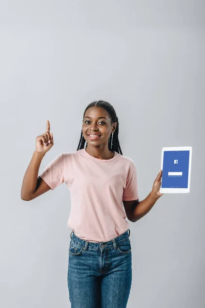 KYIV, UKRAINE - AUGUST 2, 2019: African American woman holding digital tablet with Facebook application and showing idea sign isolated on grey — Stock Photo