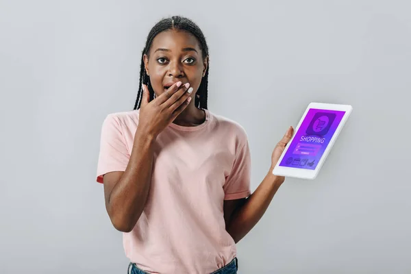 Mujer afroamericana sosteniendo tableta digital con aplicación de compras y cubriendo la boca con la mano aislada en gris - foto de stock