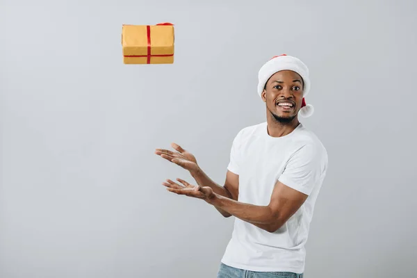 African American man in Santa hat throwing up gift box isolated on grey — Stock Photo