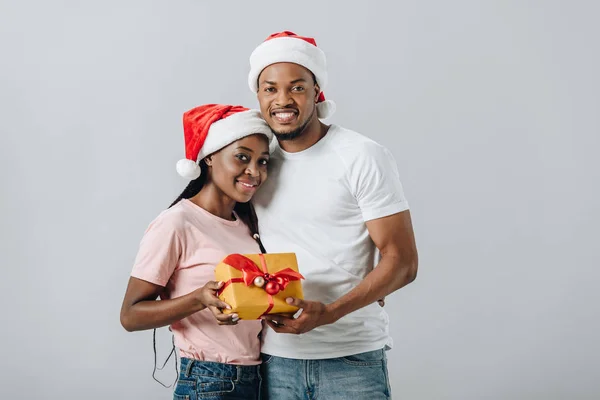 Africano casal americano em Santa chapéu abraçando e segurando caixa de presente isolado em cinza — Fotografia de Stock