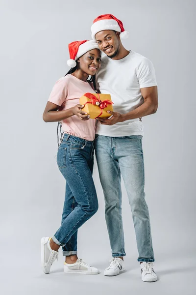 Coppia afroamericana in cappello Babbo Natale guardando la fotocamera, abbracciando e tenendo la scatola regalo isolata sul grigio — Foto stock