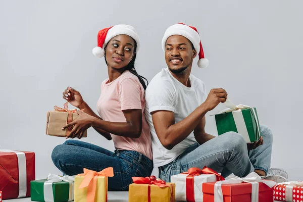 Afrikanisches amerikanisches Paar mit Weihnachtsmütze, Rücken an Rücken sitzend, Geschenke öffnend und einander isoliert auf grau betrachtend — Stockfoto