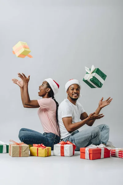 African American couple in Santa hat sitting back to back and throwing up gift boxes isolated on grey — Stock Photo