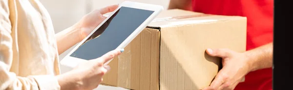 Panoramic shot of delivery man holding package near woman with digital tablet — Stock Photo