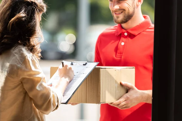 Vue recadrée de heureux livreur homme tenant boîte près de la femme avec presse-papiers et stylo — Photo de stock