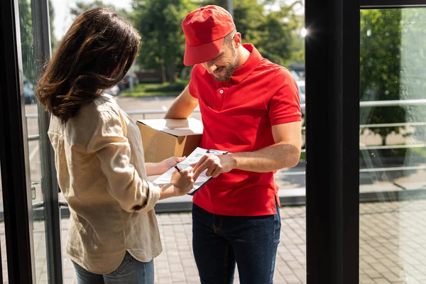 Delivery man in cap holding box and gesturing near clipboard and woman — Stock Photo