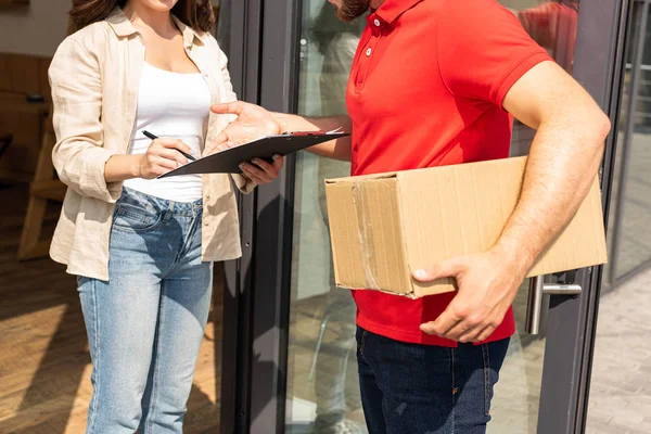 Recortado vista de entrega hombre celebración caja cerca de chica con portapapeles - foto de stock