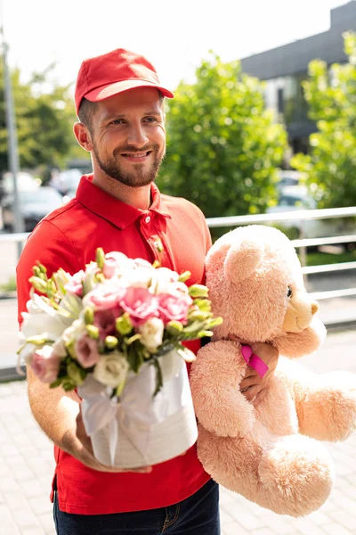 Enfoque selectivo de entrega alegre hombre sosteniendo oso de peluche y flores - foto de stock