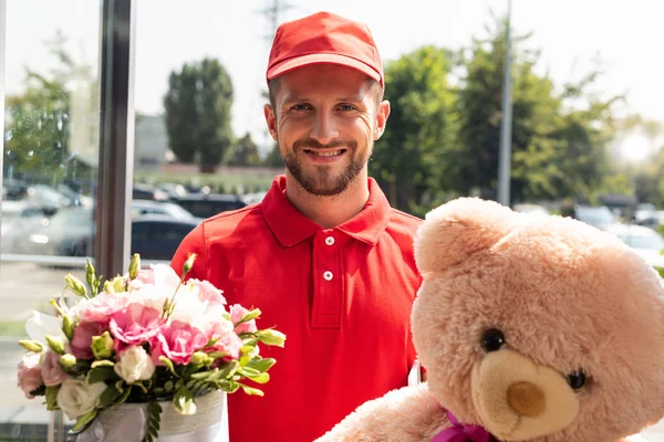 Homem de entrega sorridente em cap segurando ursinho de pelúcia e flores — Fotografia de Stock
