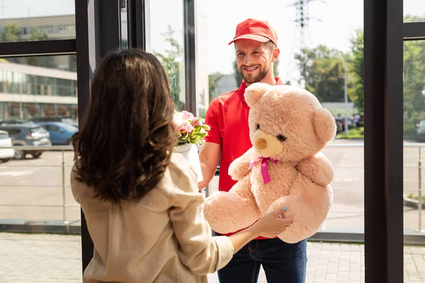Selective focus of happy delivery man in cap holding teddy bear and flowers near woman — Stock Photo