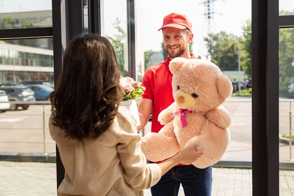 Messa a fuoco selettiva di felice consegna uomo in cap tenendo orsacchiotto e fiori vicino alla donna — Foto stock