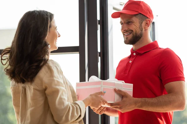 Schöner Liefermann mit Mütze schenkt glücklicher Frau ein rosafarbenes Geschenk — Stockfoto