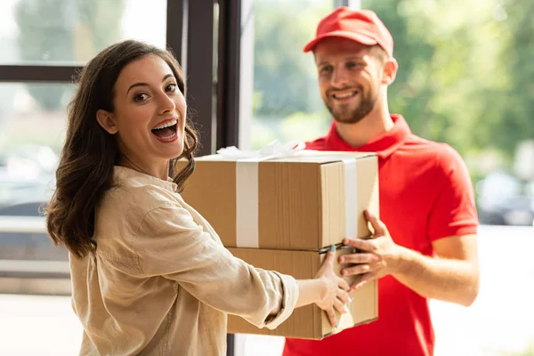 Selective focus of happy woman receiving carton boxes from cheerful delivery man in cap — Stock Photo