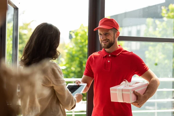 Selective focus of happy delivery man holding pink present and giving digital tablet to woman — Stock Photo