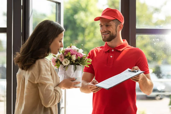 Bearded and happy delivery man holding clipboard near beautiful woman smelling flowers — Stock Photo