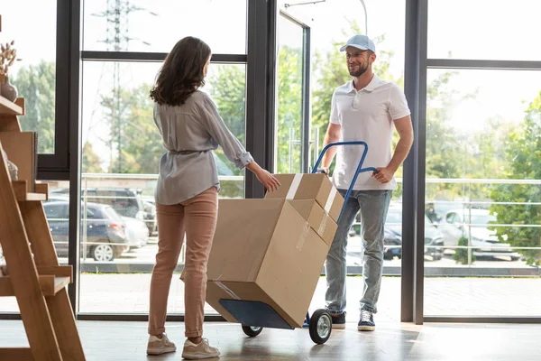 Beau livreur tenant chariot de livraison avec des boîtes près de la femme — Photo de stock