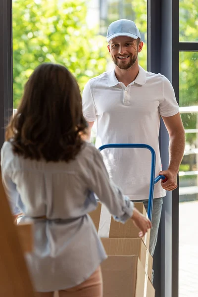 Selective focus of happy delivery man holding delivery cart with boxes near woman — Stock Photo