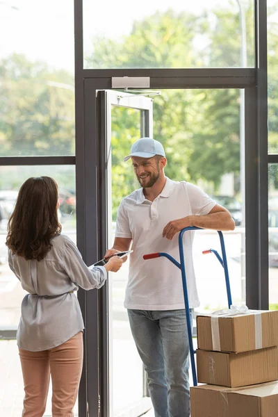 Felice uomo di consegna in piedi vicino al carrello di consegna con scatole e donna con tablet digitale — Foto stock