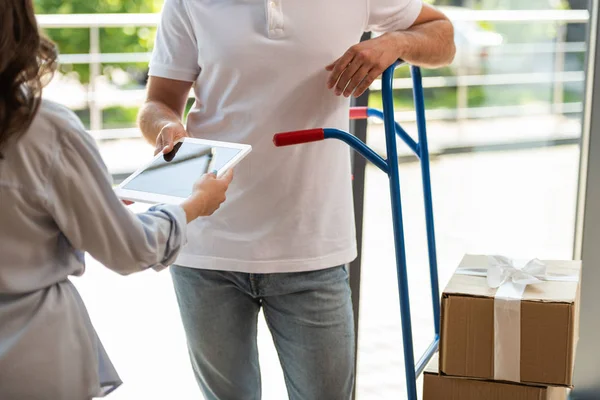 Vue recadrée du livreur debout près du chariot de livraison avec boîtes et femme avec tablette numérique — Photo de stock