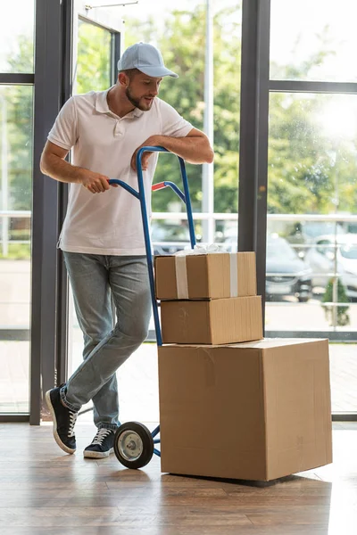 Handsome delivery man in cap standing near delivery cart and looking at cardboard boxes — Stock Photo
