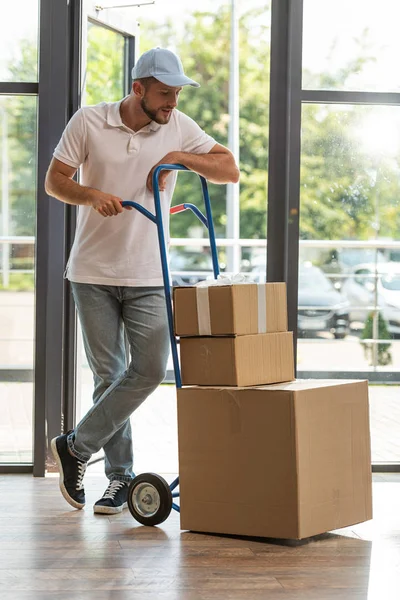 Hombre de entrega guapo en la tapa de pie cerca del carrito de entrega y mirando cajas de cartón - foto de stock