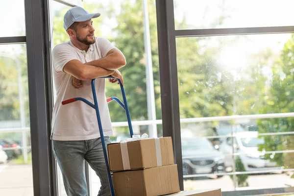 Handsome delivery man standing near delivery cart with cardboard boxes — Stock Photo