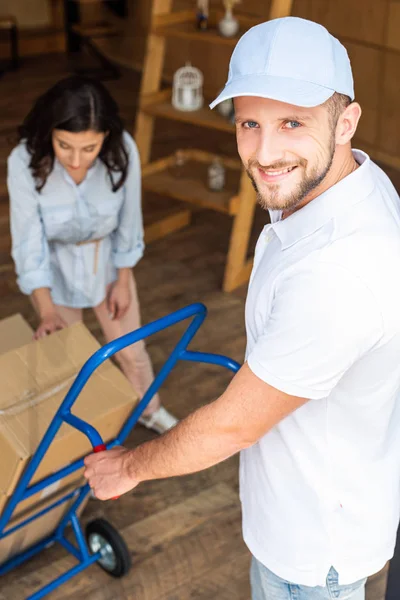 Selective focus of handsome delivery man in cap holding delivery cart near woman — Stock Photo